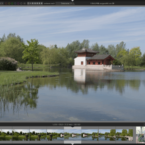 Ein Pavillon im traditionellen Stil steht an einem spiegelnden Teich, der von Bäumen umgeben ist, mit blauem Himmel und Wolkenfetzen über dem Kopf, dargestellt in einer Fotobearbeitungssoftware. (KI generierte Beschreibung)