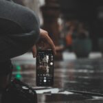Eine Person hockt auf einem nassen Bürgersteig und hält ein Smartphone in der Hand, um ein Foto von einem Gebäude zu machen, das sich in einer Pfütze spiegelt. (KI generierte Beschreibung)