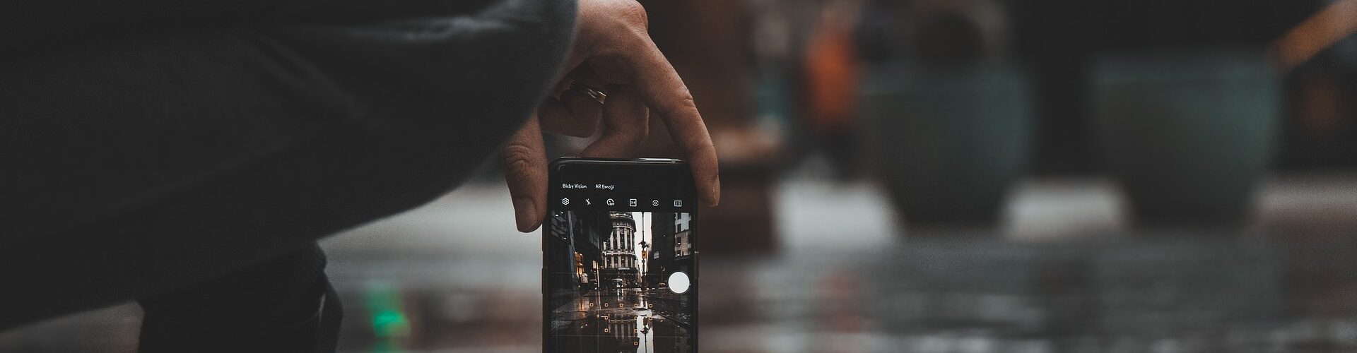 Eine Person hockt auf einem nassen Bürgersteig und hält ein Smartphone in der Hand, um ein Foto von einem Gebäude zu machen, das sich in einer Pfütze spiegelt. (KI generierte Beschreibung)