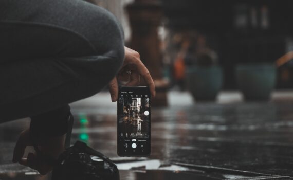 Eine Person hockt auf einem nassen Bürgersteig und hält ein Smartphone in der Hand, um ein Foto von einem Gebäude zu machen, das sich in einer Pfütze spiegelt. (KI generierte Beschreibung)