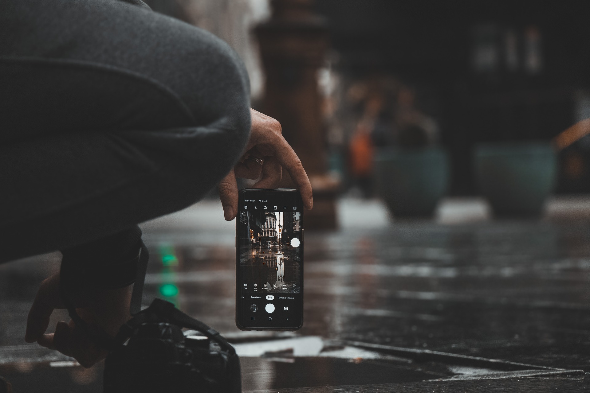 Eine Person hockt auf einem nassen Bürgersteig und hält ein Smartphone in der Hand, um ein Foto von einem Gebäude zu machen, das sich in einer Pfütze spiegelt. (KI generierte Beschreibung)