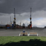 Eine Person sitzt allein auf einer Bank mit Blick auf einen Hafen mit Kränen und einem Schiff, unter einem bewölkten Himmel am Wasser. (KI generierte Beschreibung)