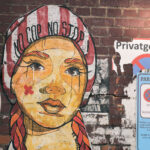 Ein Wandgemälde mit dem Gesicht einer Frau und dem Text "No Cap No Stop" ist auf eine Backsteinmauer neben Privatgrundstücken und Parkverbotsschildern in deutscher Sprache gemalt. (KI generierte Beschreibung)