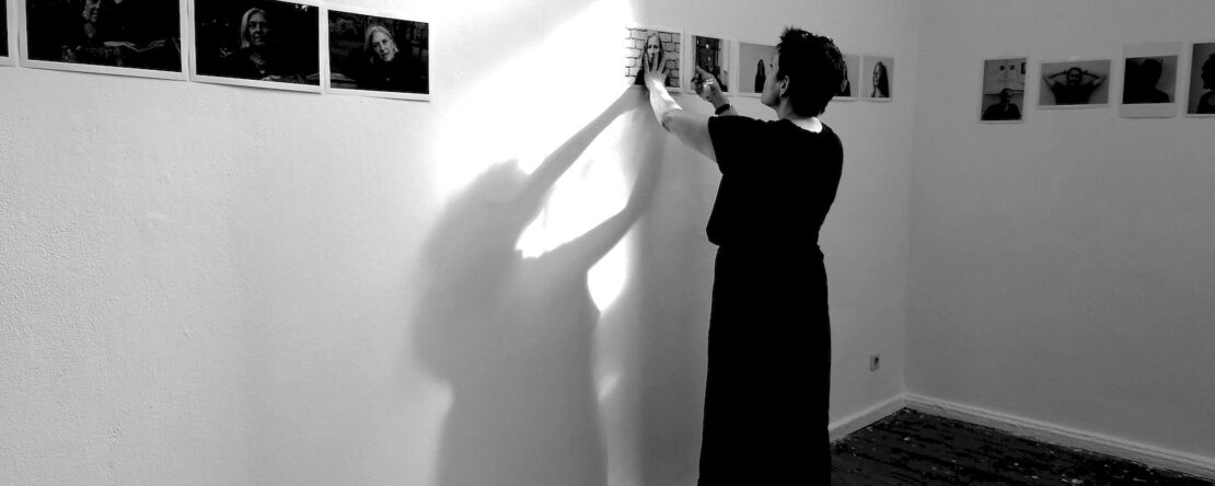 Eine Person in einem langen Kleid hängt Fotos an eine weiße Wand in einem Galerieraum und wirft einen Schatten; weitere Fotos sind entlang der Wand zu sehen. (KI generierte Beschreibung)