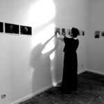 Eine Person in einem langen Kleid hängt Fotos an eine weiße Wand in einem Galerieraum und wirft einen Schatten; weitere Fotos sind entlang der Wand zu sehen. (KI generierte Beschreibung)