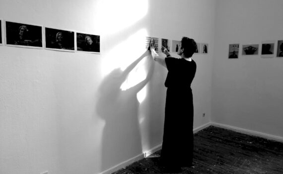 Eine Person in einem langen Kleid hängt Fotos an eine weiße Wand in einem Galerieraum und wirft einen Schatten; weitere Fotos sind entlang der Wand zu sehen. (KI generierte Beschreibung)