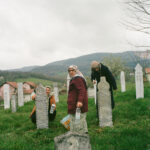 Drei Frauen säubern und pflegen Gräber auf einem grasbewachsenen Friedhof mit Plastikflaschen und Tüchern, im Hintergrund sind Häuser und Berge unter einem bewölkten Himmel zu sehen. (KI generierte Beschreibung)