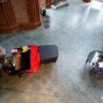 Eine Person spielt auf einem mit einem rot-schwarzen Tuch bedeckten Flügel in einem geräumigen Raum mit Backsteinwänden, einem runden Tisch und Mänteln auf einem Gestell. (KI generierte Beschreibung)