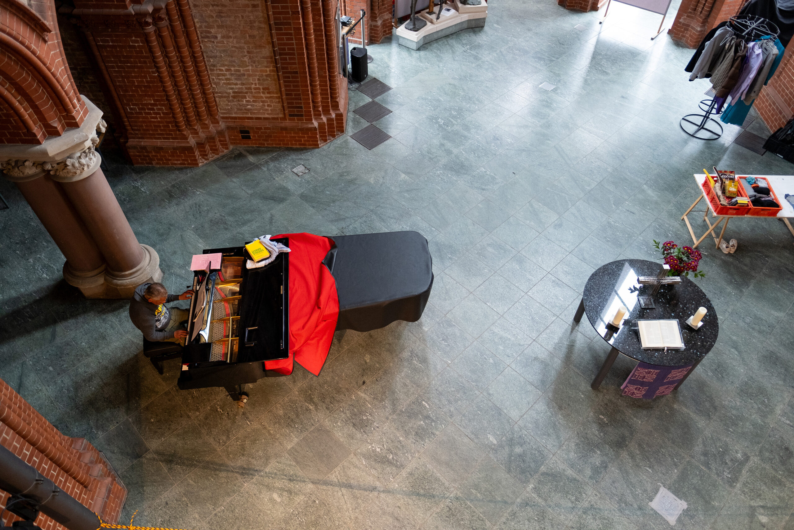 Eine Person spielt auf einem mit einem rot-schwarzen Tuch bedeckten Flügel in einem geräumigen Raum mit Backsteinwänden, einem runden Tisch und Mänteln auf einem Gestell. (KI generierte Beschreibung)