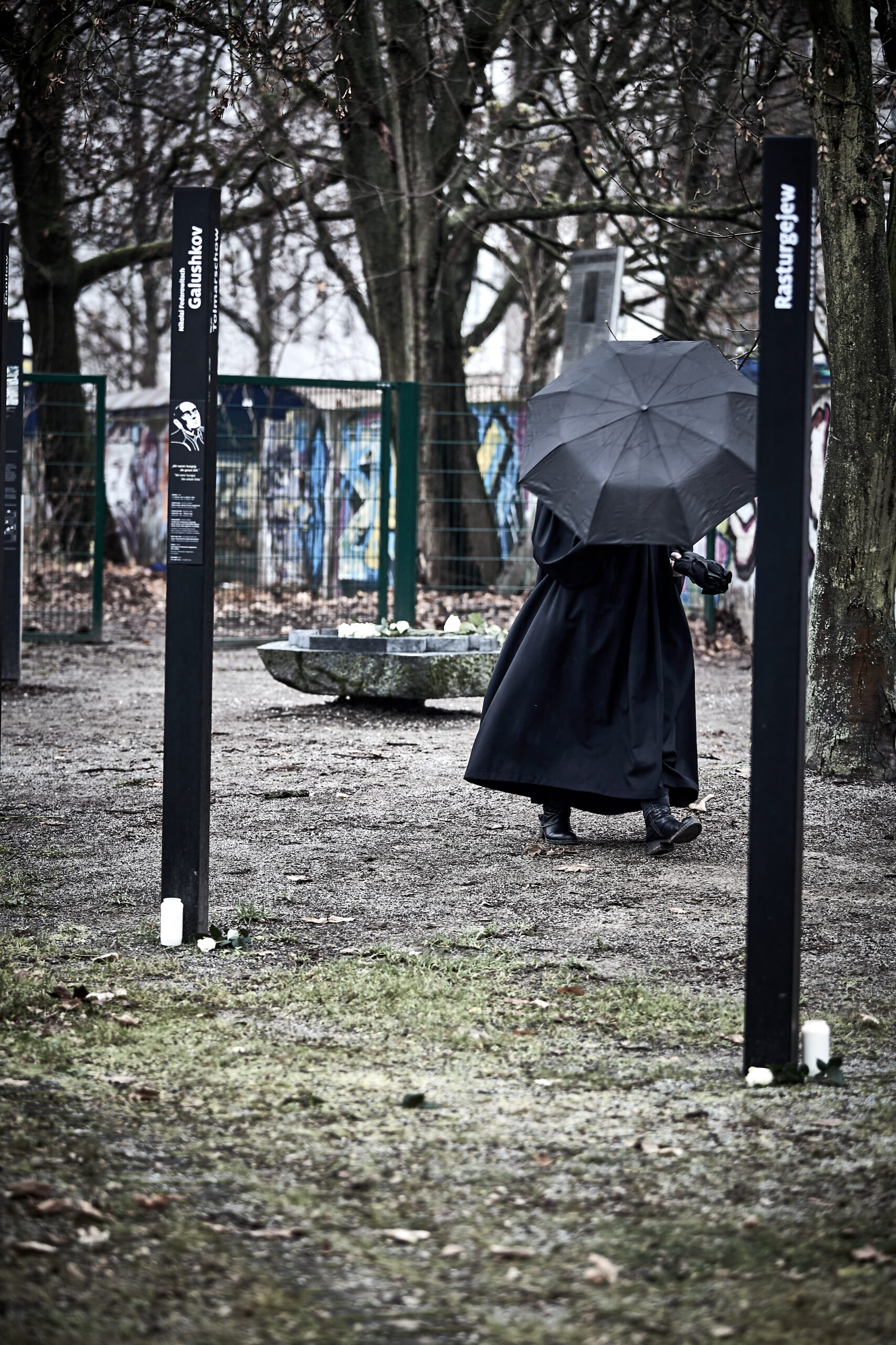 Eine Person in einem langen dunklen Mantel geht mit einem großen schwarzen Regenschirm durch einen Außenbereich, der von Bäumen und mit Graffiti beschmierten Mauern begrenzt wird. (KI generierte Beschreibung)