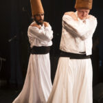 Zwei Männer in traditionellen weißen Gewändern und hohen braunen Hüten führen auf der Bühne einen wirbelnden Sufi-Tanz auf, bei dem sie die Arme vor der Brust verschränken. (KI generierte Beschreibung)