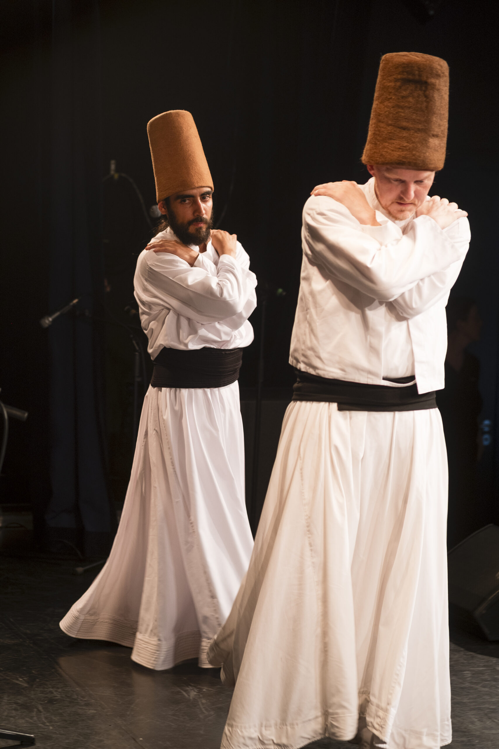 Zwei Männer in traditionellen weißen Gewändern und hohen braunen Hüten führen auf der Bühne einen wirbelnden Sufi-Tanz auf, bei dem sie die Arme vor der Brust verschränken. (KI generierte Beschreibung)
