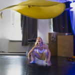 Eine Frau in Freizeitkleidung hockt auf einem schwarzen Boden in einem Innenraum und reagiert, als ein großes gelbes Tuch über ihr schwebt. (KI generierte Beschreibung)