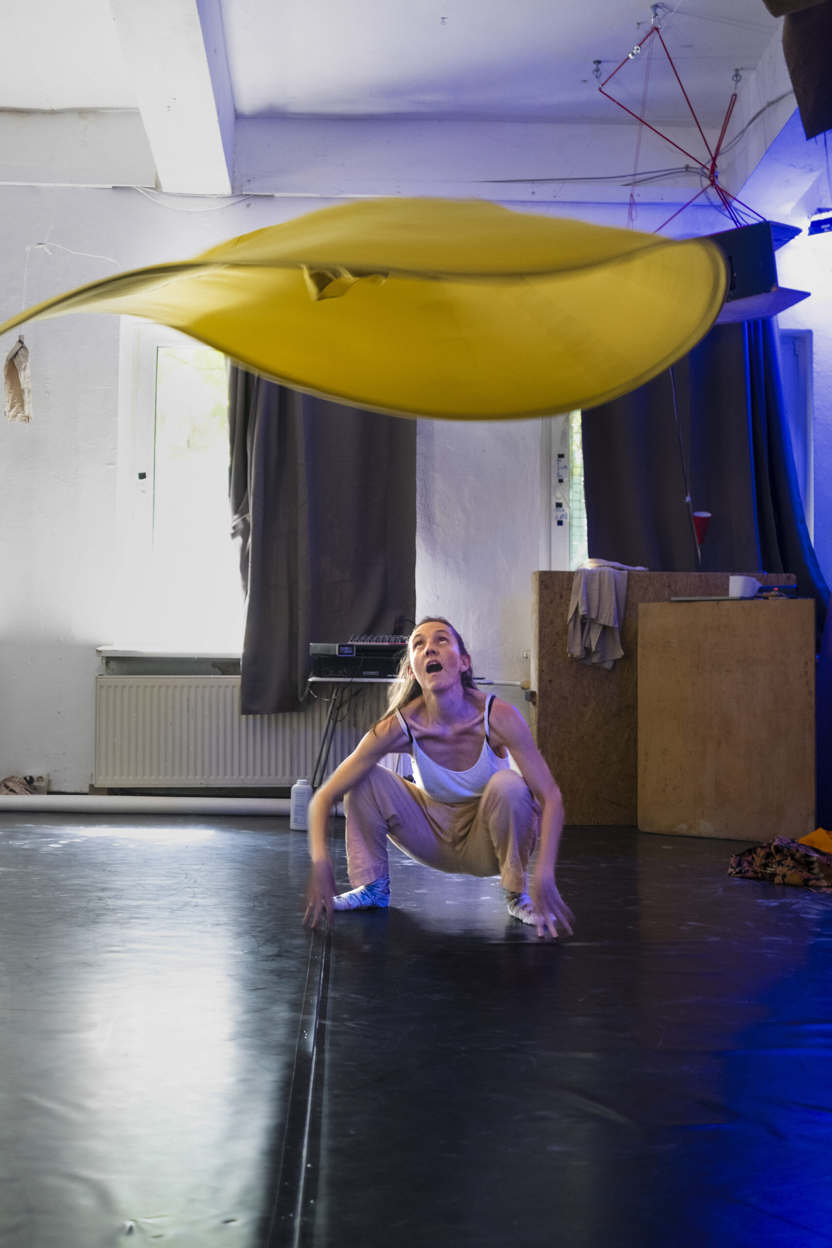 Eine Frau in Freizeitkleidung hockt auf einem schwarzen Boden in einem Innenraum und reagiert, als ein großes gelbes Tuch über ihr schwebt. (KI generierte Beschreibung)