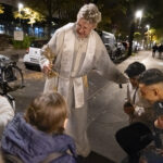 Eine Person in religiöser Kleidung hält einen großen Schlüssel und spricht zu einer Gruppe von Menschen auf einem nächtlichen Bürgersteig. (KI generierte Beschreibung)