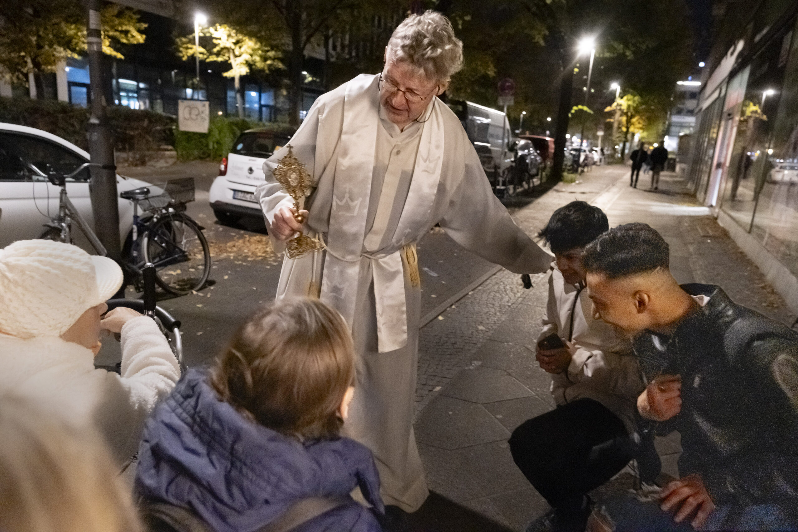 Eine Person in religiöser Kleidung hält einen großen Schlüssel und spricht zu einer Gruppe von Menschen auf einem nächtlichen Bürgersteig. (KI generierte Beschreibung)