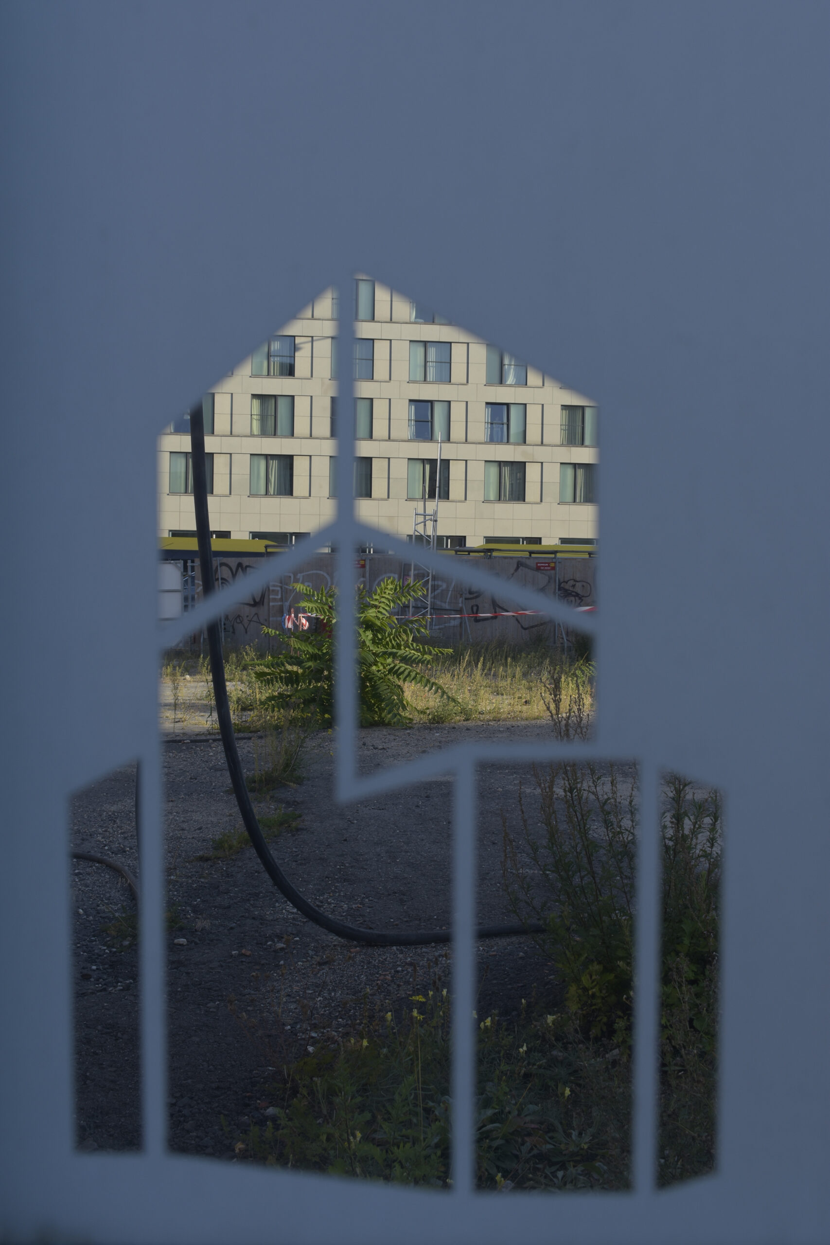 Ein Gebäude und ein bewachsenes Grundstück sind durch einen geometrischen, hausförmigen Ausschnitt im Vordergrund sichtbar. (KI generierte Beschreibung)