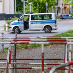 Ein deutscher Polizeiwagen steht mit offener Tür auf einer Kopfsteinpflasterstraße; ein weiteres Polizeifahrzeug ist weiter unten auf der Straße hinter rot-weißen Metallbarrieren zu sehen. (KI generierte Beschreibung)