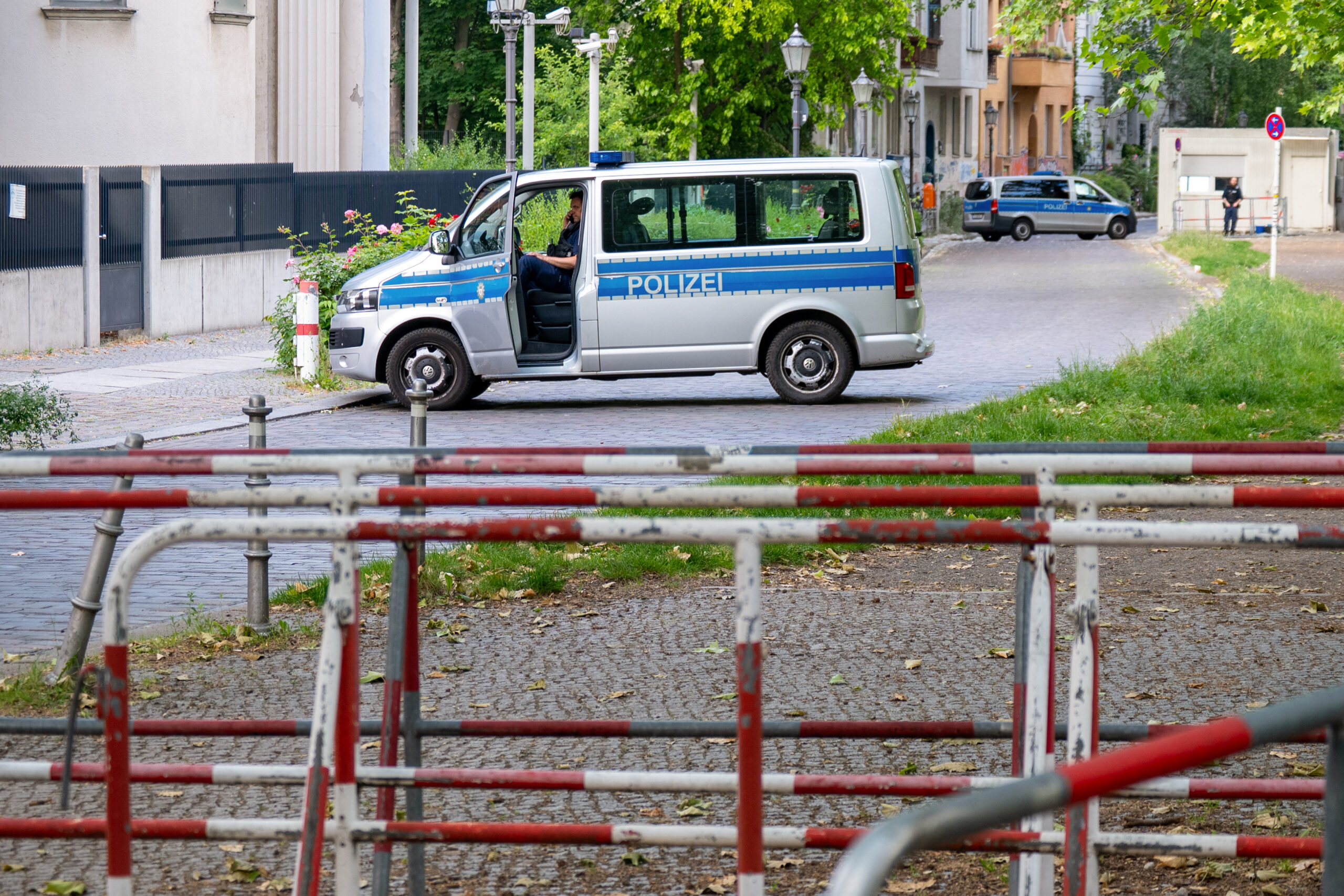 Ein deutscher Polizeiwagen steht mit offener Tür auf einer Kopfsteinpflasterstraße; ein weiteres Polizeifahrzeug ist weiter unten auf der Straße hinter rot-weißen Metallbarrieren zu sehen. (KI generierte Beschreibung)