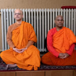 Zwei buddhistische Mönche in orangefarbenen Gewändern sitzen im Schneidersitz auf einer Plattform vor einem Heizkörper, die Augen geschlossen, und scheinen zu meditieren. Neben ihnen liegen verschiedene Gegenstände auf dem Boden. (KI generierte Beschreibung)