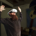 Eine bärtige Person, die ein Virtual-Reality-Headset trägt, steht mit erhobenen Armen in einem Raum mit abstrakter Kunst und verschiedenen Objekten im Hintergrund. (KI generierte Beschreibung)