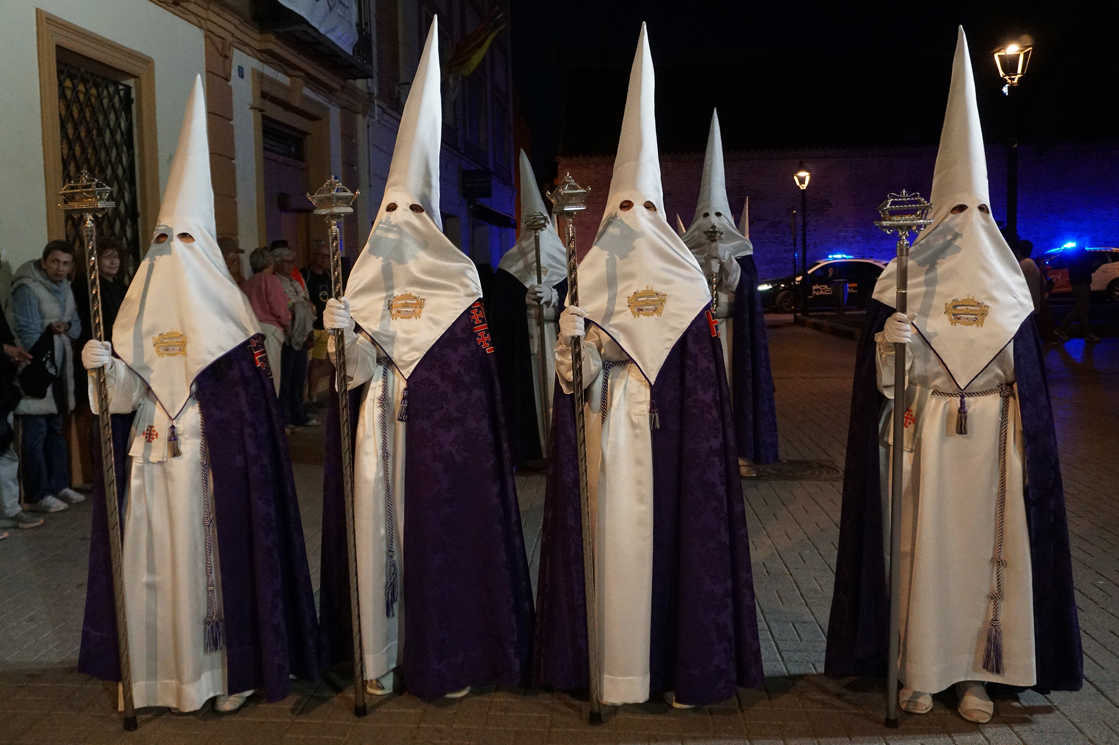 Mehrere Personen in weißen Kapuzen und violetten Gewändern stehen nachts auf einer Straße und nehmen wahrscheinlich an einer religiösen Prozession teil. (KI generierte Beschreibung)