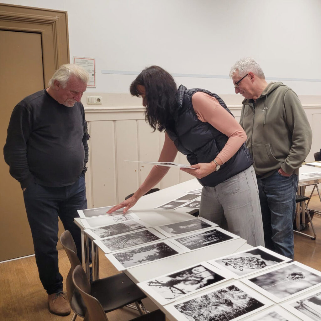 Drei Erwachsene stehen um einen mit Schwarz-Weiß-Fotos bedeckten Tisch; eine Frau zeigt auf ein Foto, während die beiden Männer es betrachten. (KI generierte Beschreibung)