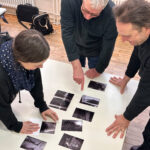 Drei Personen stehen um einen Tisch herum und betrachten und diskutieren mehrere Schwarz-Weiß-Fotos, die in einem Raster angeordnet sind. (KI generierte Beschreibung)