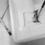 Mit einem Pinsel wird ein dünner Film auf das Wasser in einer Schale aufgetragen, während eine Schere und ein weiterer Pinsel in der Nähe auf der Oberfläche liegen. (KI generierte Beschreibung)