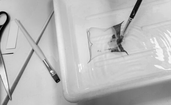 Mit einem Pinsel wird ein dünner Film auf das Wasser in einer Schale aufgetragen, während eine Schere und ein weiterer Pinsel in der Nähe auf der Oberfläche liegen. (KI generierte Beschreibung)