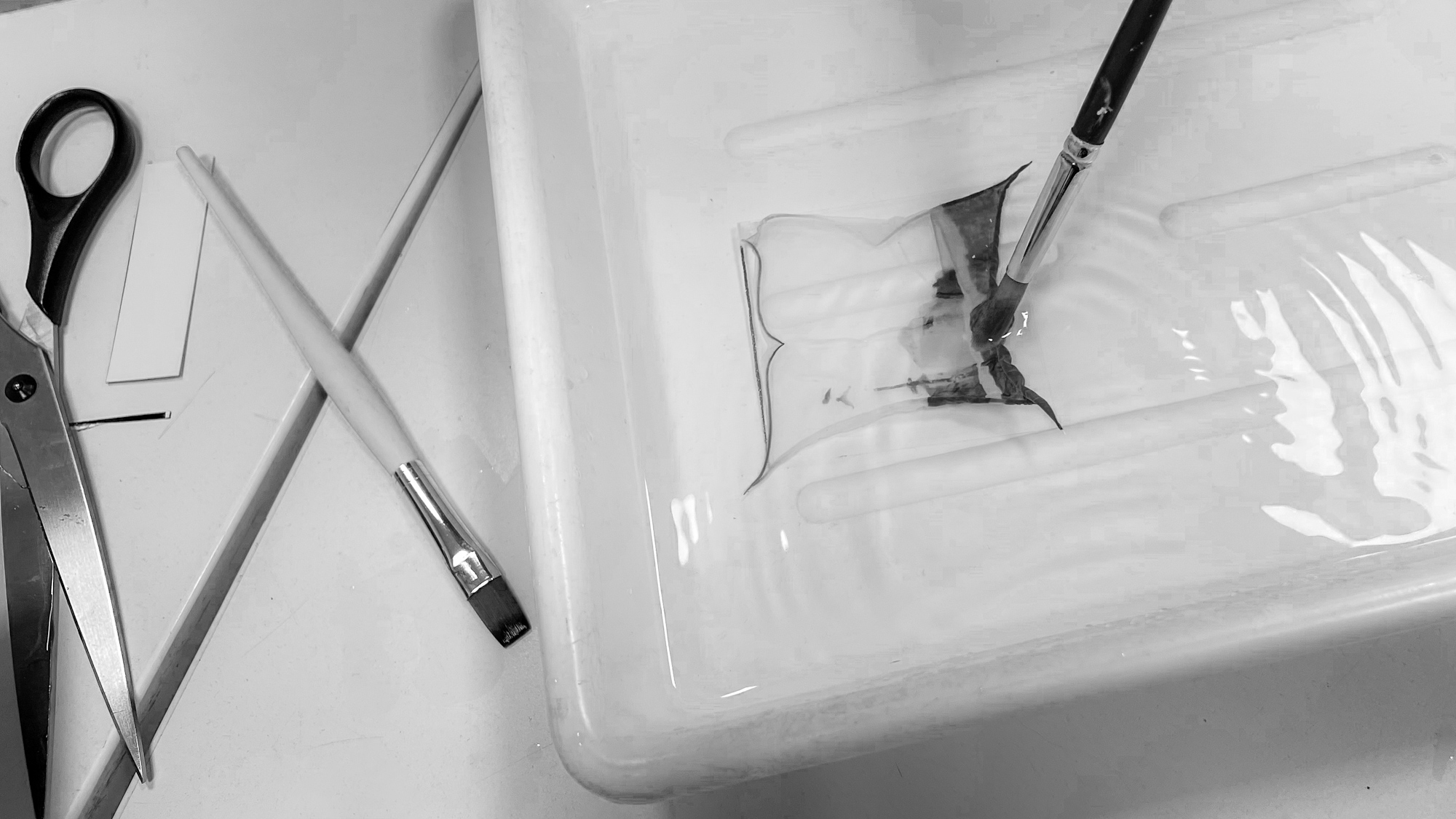 Mit einem Pinsel wird ein dünner Film auf das Wasser in einer Schale aufgetragen, während eine Schere und ein weiterer Pinsel in der Nähe auf der Oberfläche liegen. (KI generierte Beschreibung)