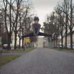 Eine Person springt in einem Spagat mitten in der Luft über einen gepflasterten Weg, während eine ältere Person auf der rechten Seite steht und eine Papiertüte hält. Bäume und Gebäude säumen die Straße. (KI generierte Beschreibung)