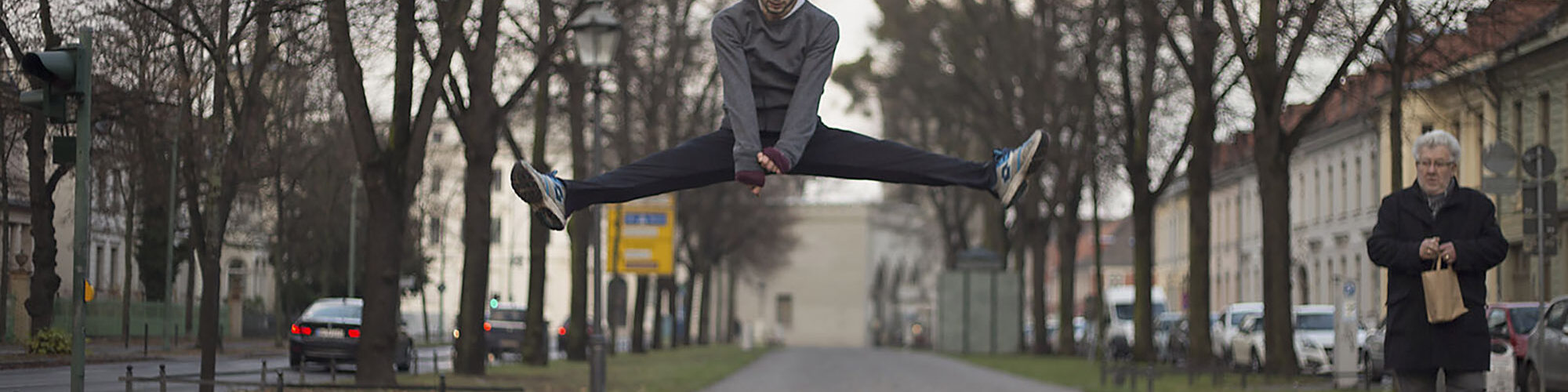Eine Person springt in einem Spagat mitten in der Luft über einen gepflasterten Weg, während eine ältere Person auf der rechten Seite steht und eine Papiertüte hält. Bäume und Gebäude säumen die Straße. (KI generierte Beschreibung)