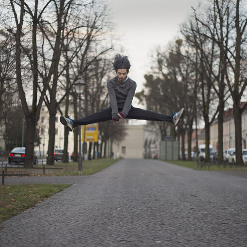 Eine Person springt in einem Spagat mitten in der Luft über einen gepflasterten Weg, während eine ältere Person auf der rechten Seite steht und eine Papiertüte hält. Bäume und Gebäude säumen die Straße. (KI generierte Beschreibung)