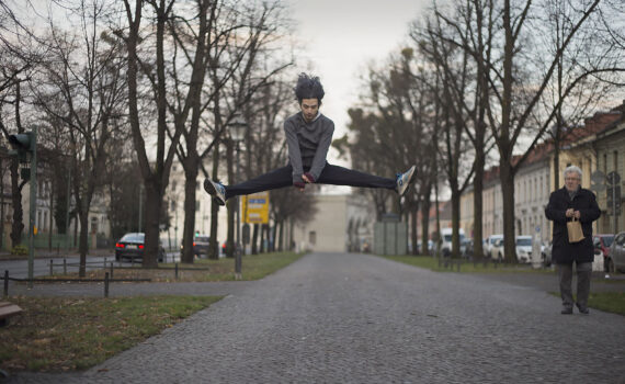 Eine Person springt in einem Spagat mitten in der Luft über einen gepflasterten Weg, während eine ältere Person auf der rechten Seite steht und eine Papiertüte hält. Bäume und Gebäude säumen die Straße. (KI generierte Beschreibung)