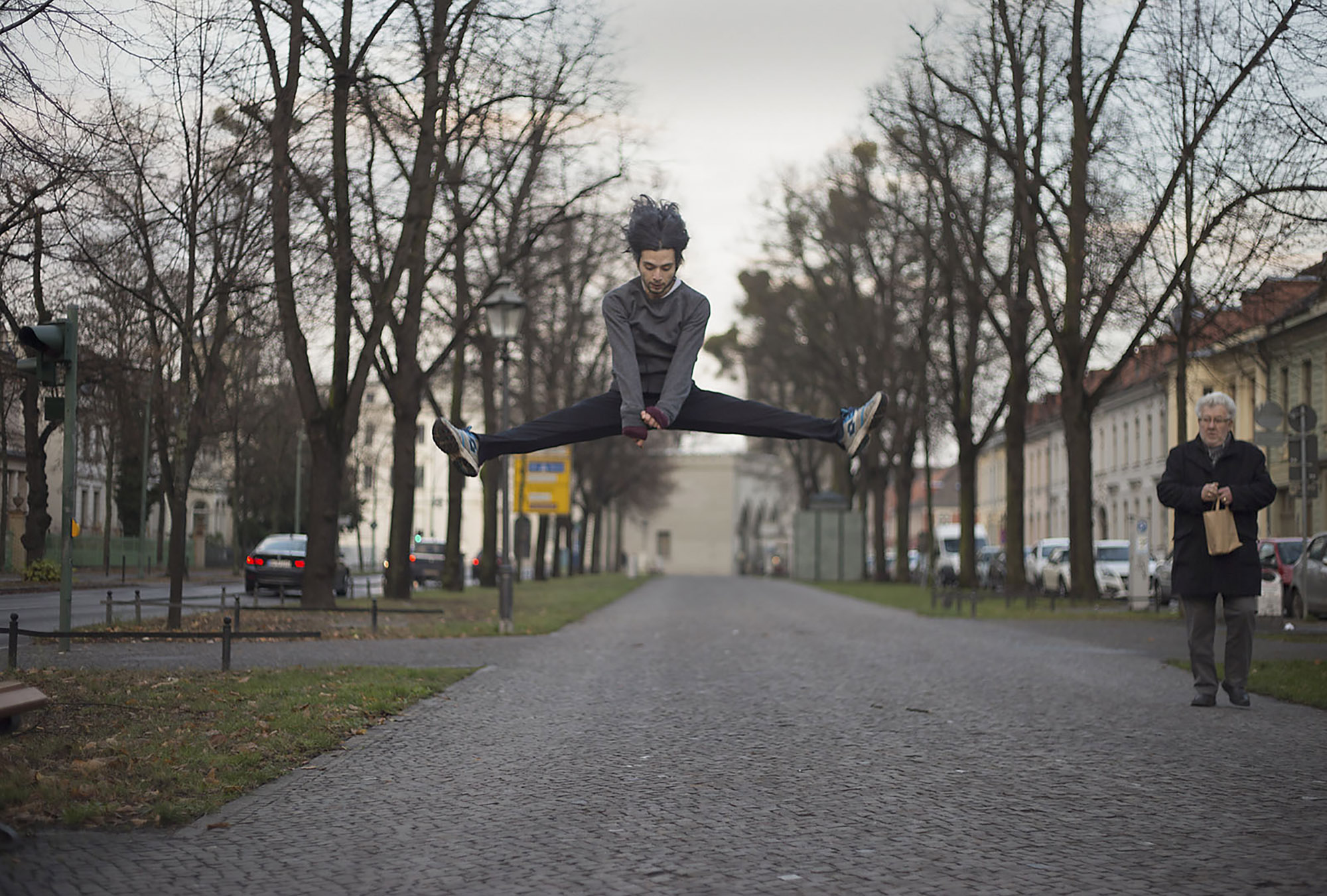 Eine Person springt in einem Spagat mitten in der Luft über einen gepflasterten Weg, während eine ältere Person auf der rechten Seite steht und eine Papiertüte hält. Bäume und Gebäude säumen die Straße. (KI generierte Beschreibung)