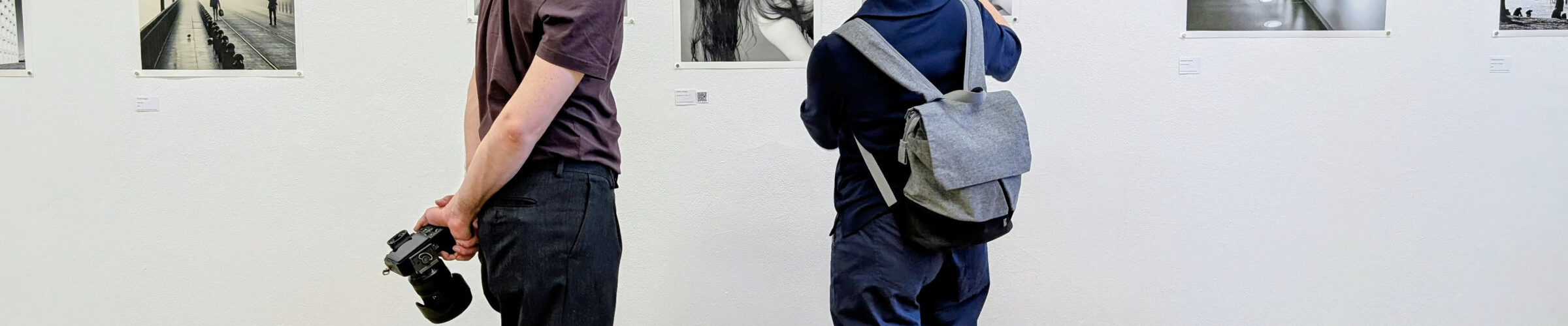 Zwei Menschen stehen in einer Kunstgalerie und betrachten Schwarz-Weiß-Fotografien an einer weißen Wand; einer hält eine Kamera hinter seinem Rücken. (KI generierte Beschreibung)