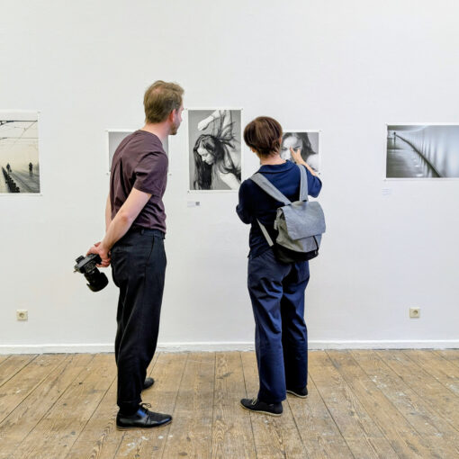 Zwei Menschen stehen in einer Kunstgalerie und betrachten Schwarz-Weiß-Fotografien an einer weißen Wand; einer hält eine Kamera hinter seinem Rücken. (KI generierte Beschreibung)