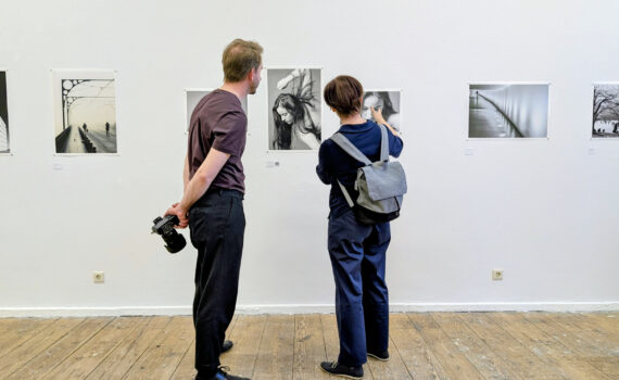 Zwei Menschen stehen in einer Kunstgalerie und betrachten Schwarz-Weiß-Fotografien an einer weißen Wand; einer hält eine Kamera hinter seinem Rücken. (KI generierte Beschreibung)