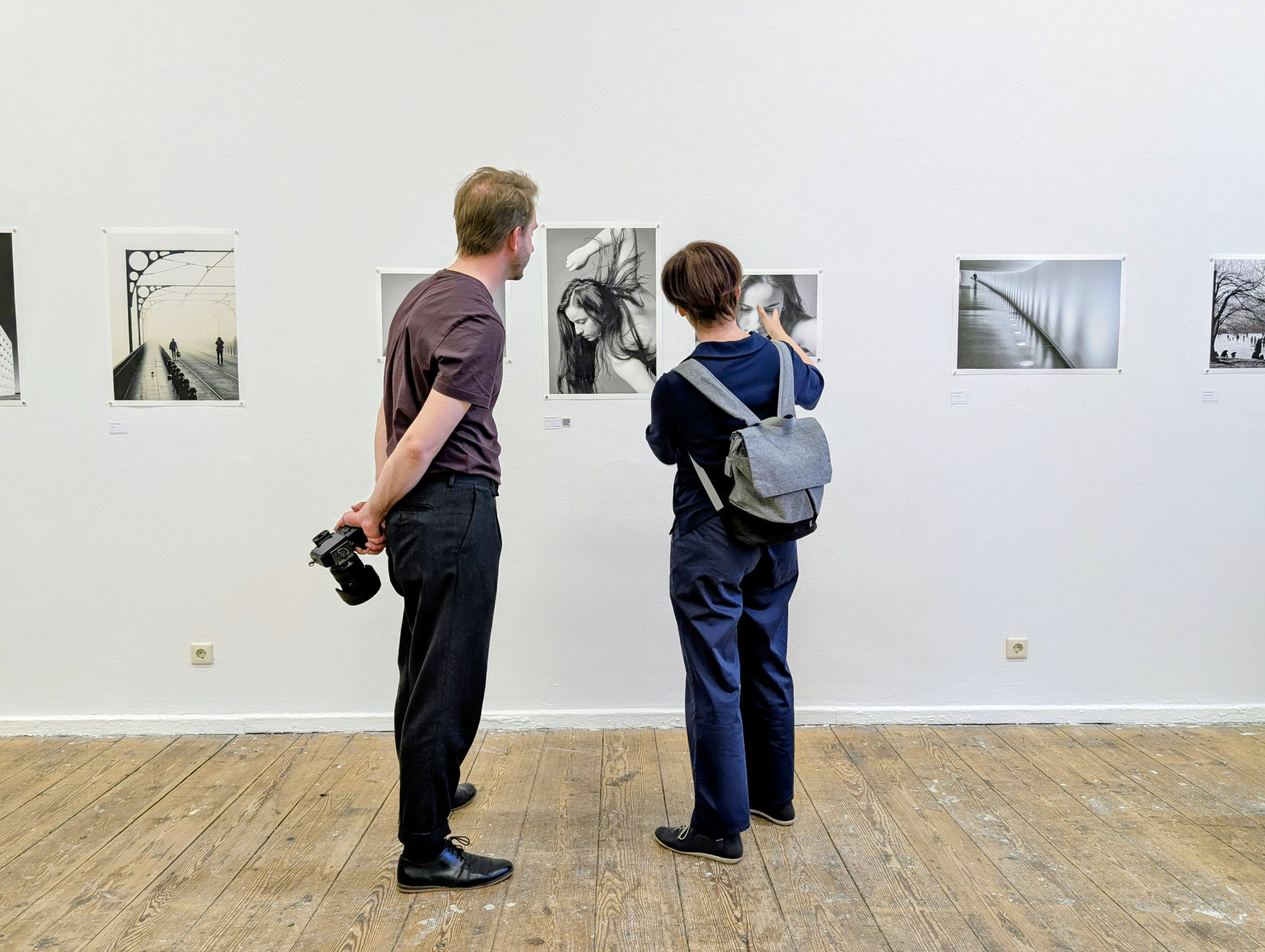 Zwei Menschen stehen in einer Kunstgalerie und betrachten Schwarz-Weiß-Fotografien an einer weißen Wand; einer hält eine Kamera hinter seinem Rücken. (KI generierte Beschreibung)