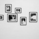 Sechs Schwarz-Weiß-Fotografien in schwarzen Rahmen sind versetzt an einer schlichten weißen Wand angeordnet. (KI generierte Beschreibung)