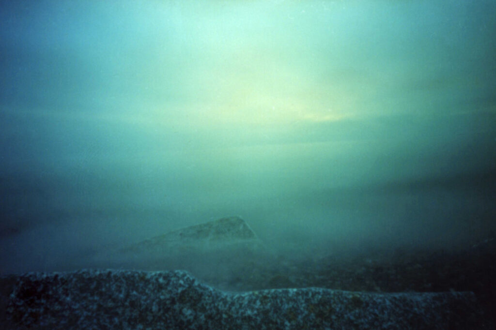 Eine neblige Meereslandschaft mit teilweise sichtbaren Felsen im Vordergrund und diffusem Licht am bewölkten Himmel. (KI generierte Beschreibung)