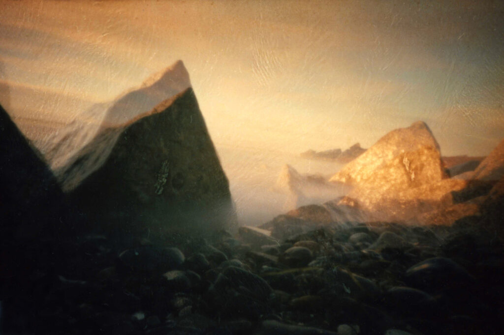 Große Felsen und Kieselsteine am Ufer werden von warmem, sanftem Sonnenlicht beleuchtet, während Nebel oder Dunst eine traumhafte, ätherische Atmosphäre schaffen. (KI generierte Beschreibung)