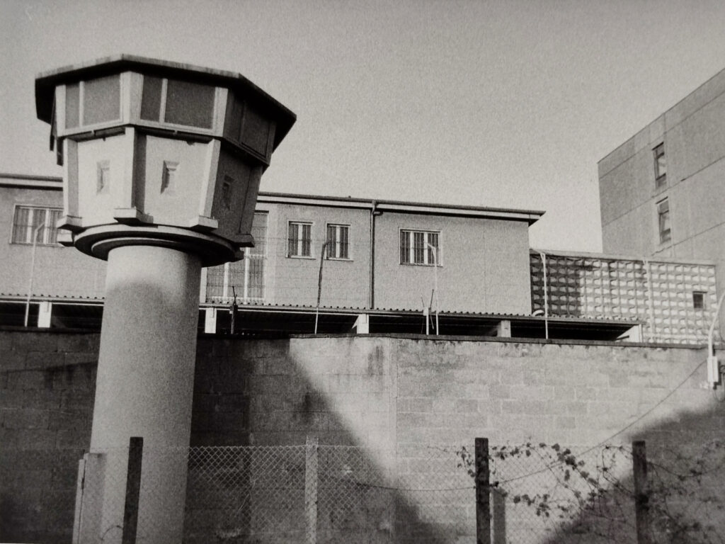 Schwarz-Weiß-Foto eines Gefängnisses oder einer Haftanstalt mit einem Wachturm, einem Stacheldrahtzaun und Betongebäuden. (KI generierte Beschreibung)