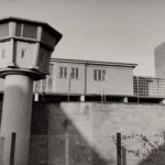 Schwarz-Weiß-Foto eines Gefängnisses oder einer Haftanstalt mit einem Wachturm, einem Stacheldrahtzaun und Betongebäuden. (KI generierte Beschreibung)