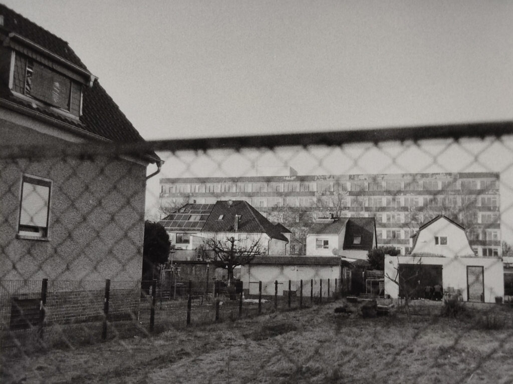 Schwarz-Weiß-Foto, auf dem im Vordergrund mehrere Häuser und ein umzäunter Hof zu sehen sind, während im Hintergrund durch einen Maschendrahtzaun große Mehrfamilienhäuser zu erkennen sind. (KI generierte Beschreibung)