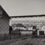 Schwarz-Weiß-Foto, auf dem im Vordergrund mehrere Häuser und ein umzäunter Hof zu sehen sind, während im Hintergrund durch einen Maschendrahtzaun große Mehrfamilienhäuser zu erkennen sind. (KI generierte Beschreibung)