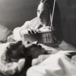 Zwei Frauen liegen auf einem Bett; die eine hält ein Radio in der Hand und schaut nach oben, während die andere mit geschlossenen Augen daliegt. Das Bild ist in Schwarz-Weiß. (KI generierte Beschreibung)
