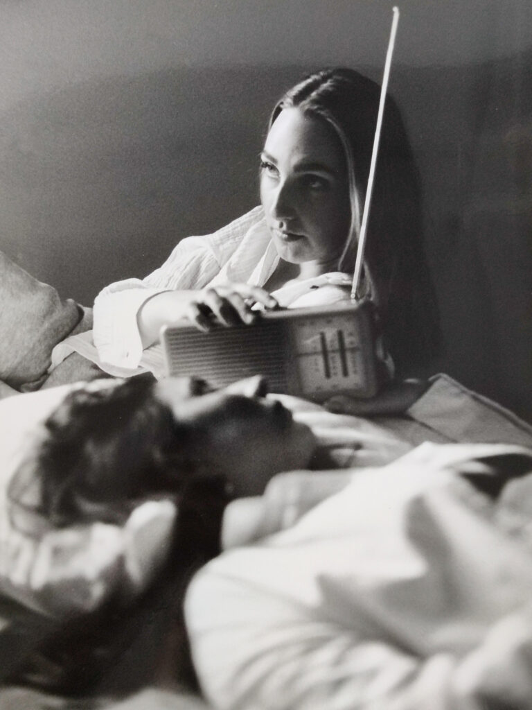 Zwei Frauen liegen auf einem Bett; die eine hält ein Radio in der Hand und schaut nach oben, während die andere mit geschlossenen Augen daliegt. Das Bild ist in Schwarz-Weiß. (KI generierte Beschreibung)