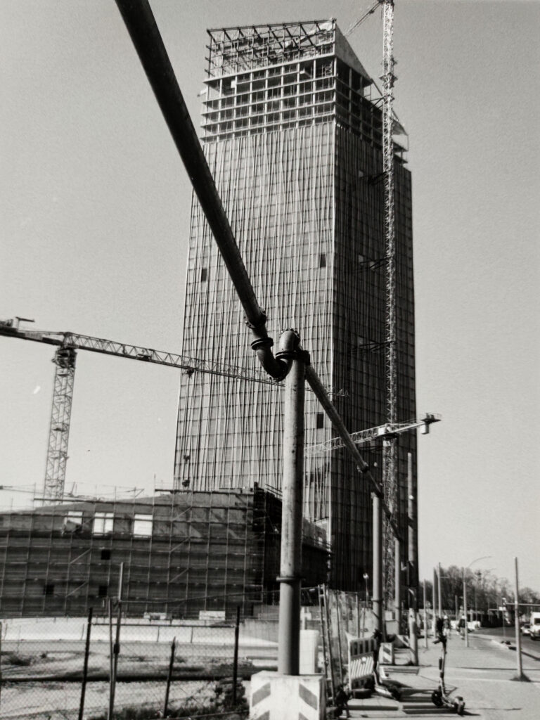 Schwarz-Weiß-Foto eines hohen Gebäudes im Bau, mit Kränen und Gerüsten rund um das Bauwerk sowie Baumaterialien im Vordergrund. (KI generierte Beschreibung)